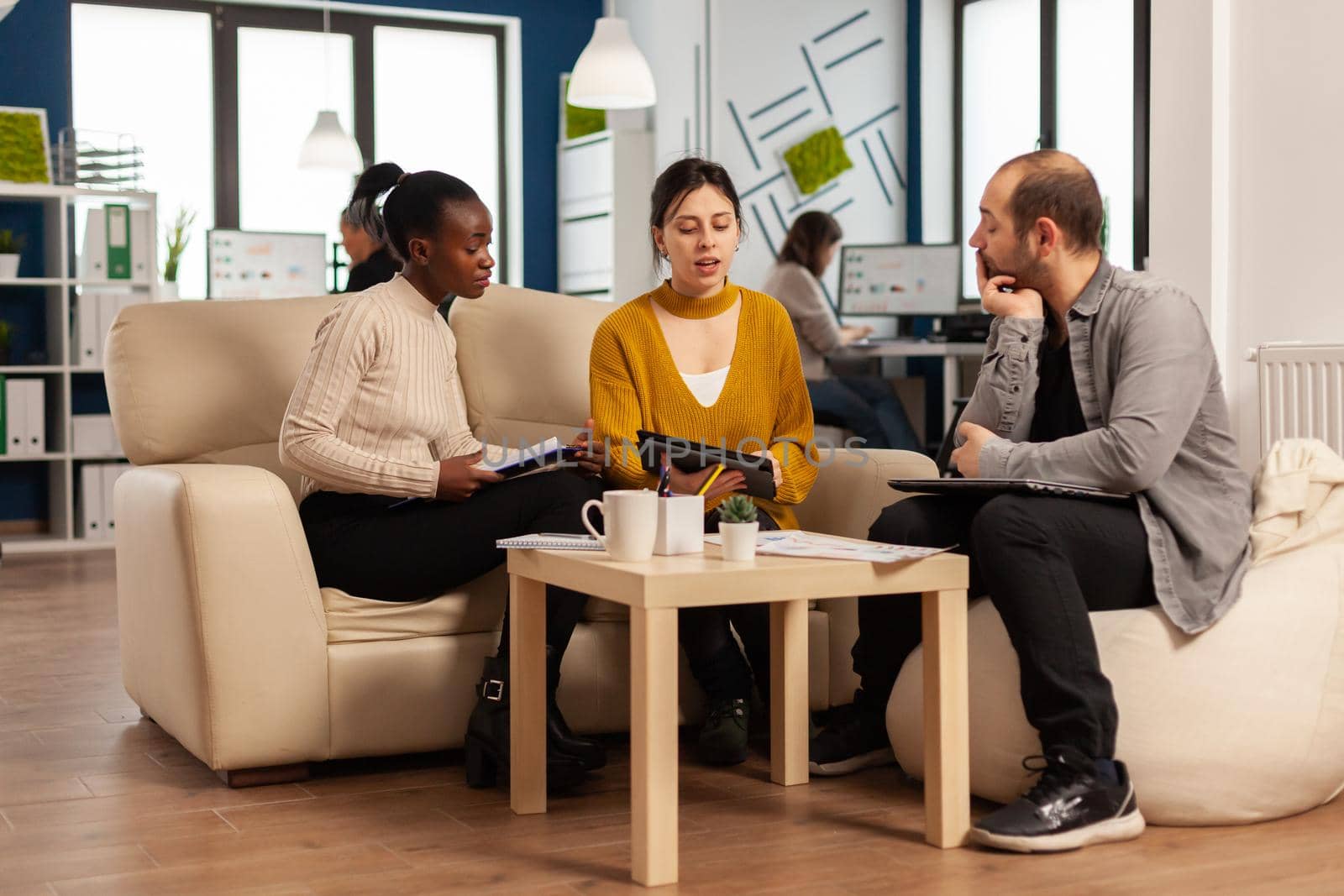 Confident company manager giving working tasks to diverse teammates sitting on couch in start up office. Multiethnic team discussing project ideas at brainstorming meeting using digital devices