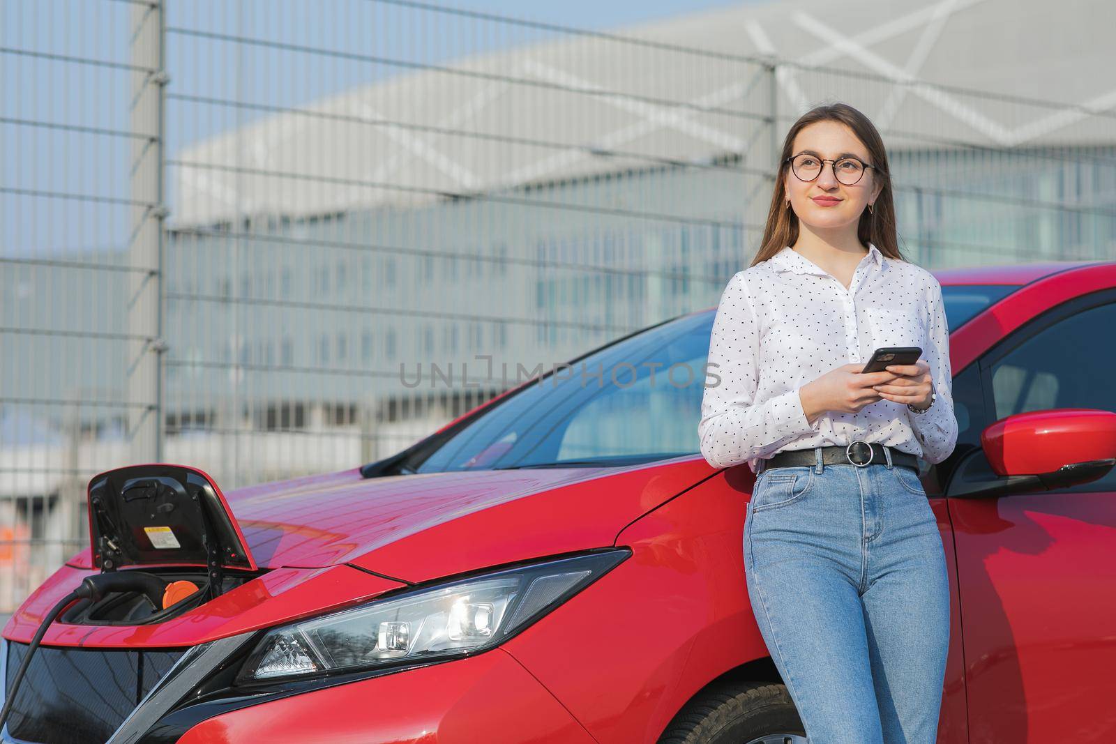 Caucasian girl using smart phone and waiting power supply connect to electric vehicles for charging the battery in car. Ecological car connected and charging batteries