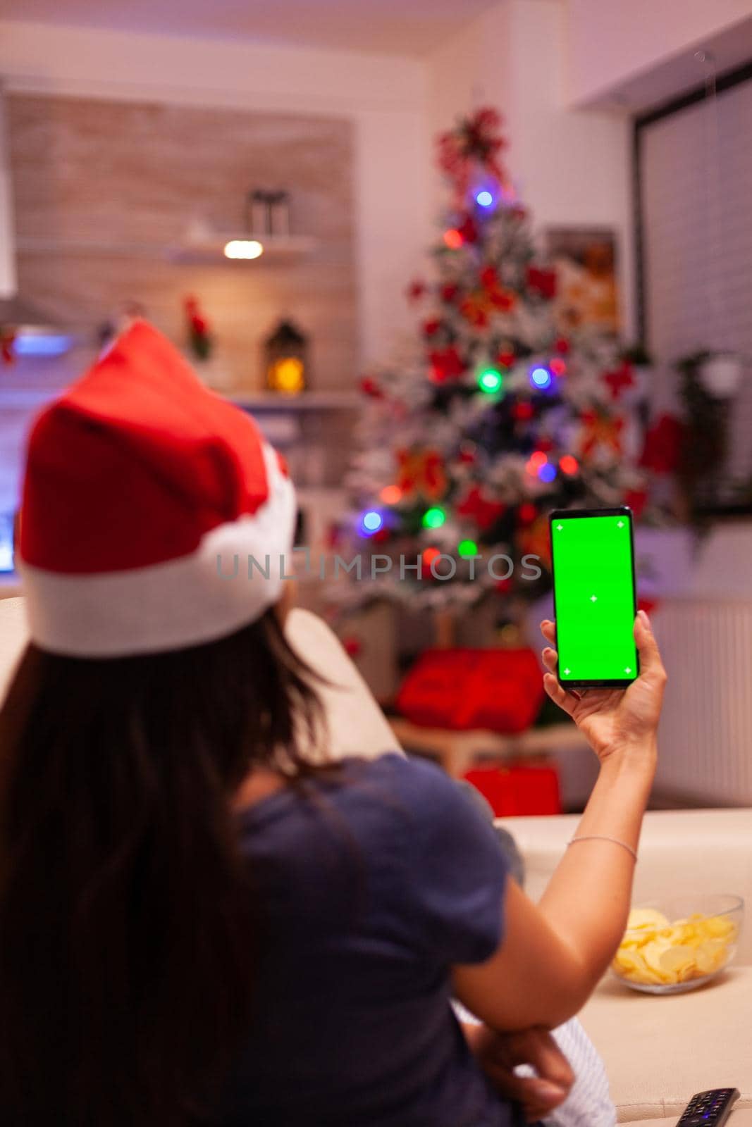Woman holding green screen mock up chroma key phone with isolated display sitting comfortable on sofa in xmas decorated kitchen. Person celebrating christmas holiday enjoying winter season