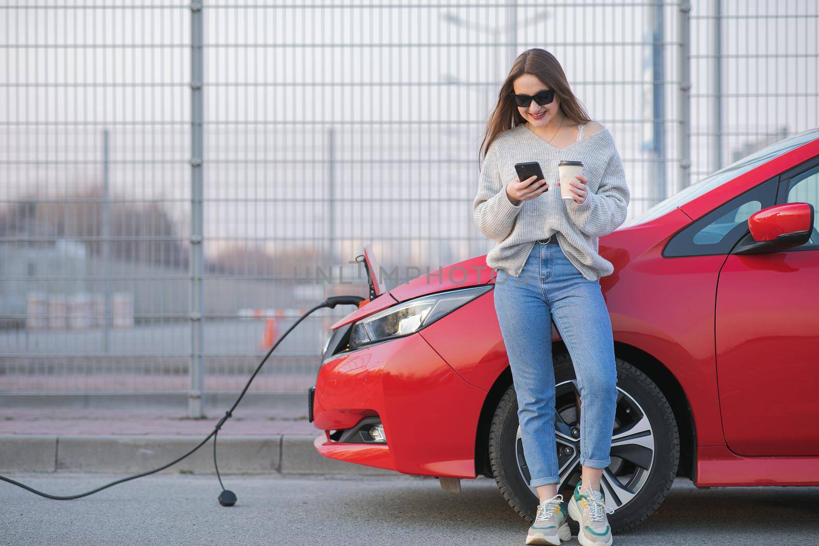 Electric car charging in street. Ecological Car Connected and Charging Batteries. Girl Use Coffee Drink While Using SmartPhone and Waiting Power Supply Connect to Electric Vehicles for Charging by uflypro