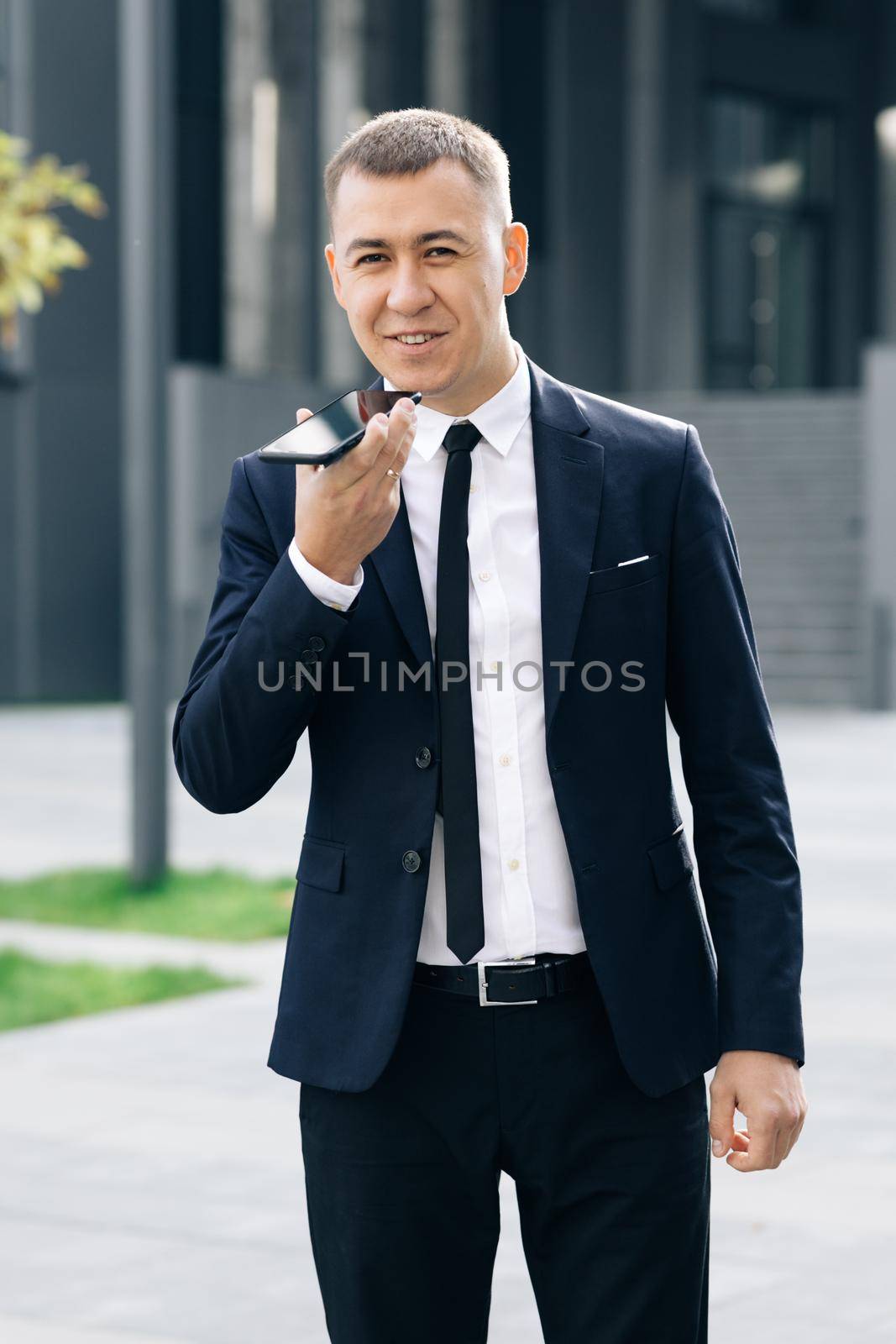 Young business man talking on phone near modern office building. Businessman use smartphone to send voice messages outdoors at downtown.