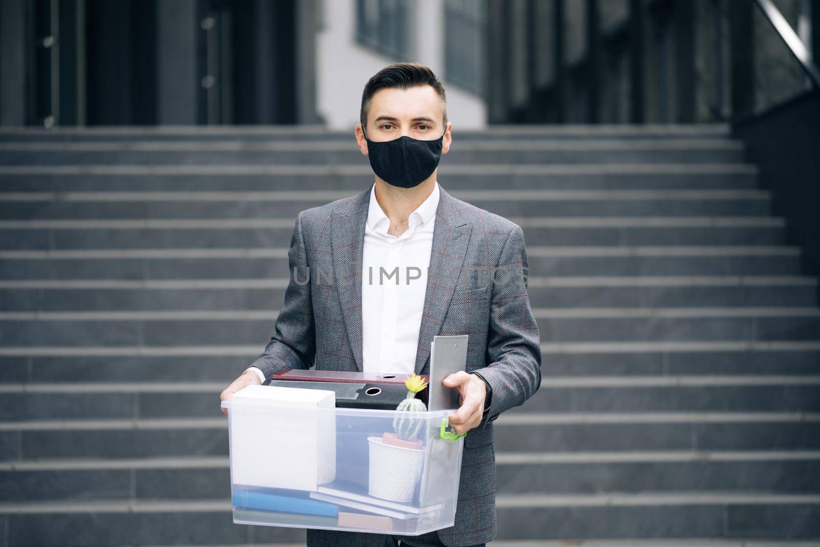 Fired sad young businessman in medical mask carries box with personal belongings leaving the office. Unemployment. Lack of jobs. Financial crisis. by uflypro