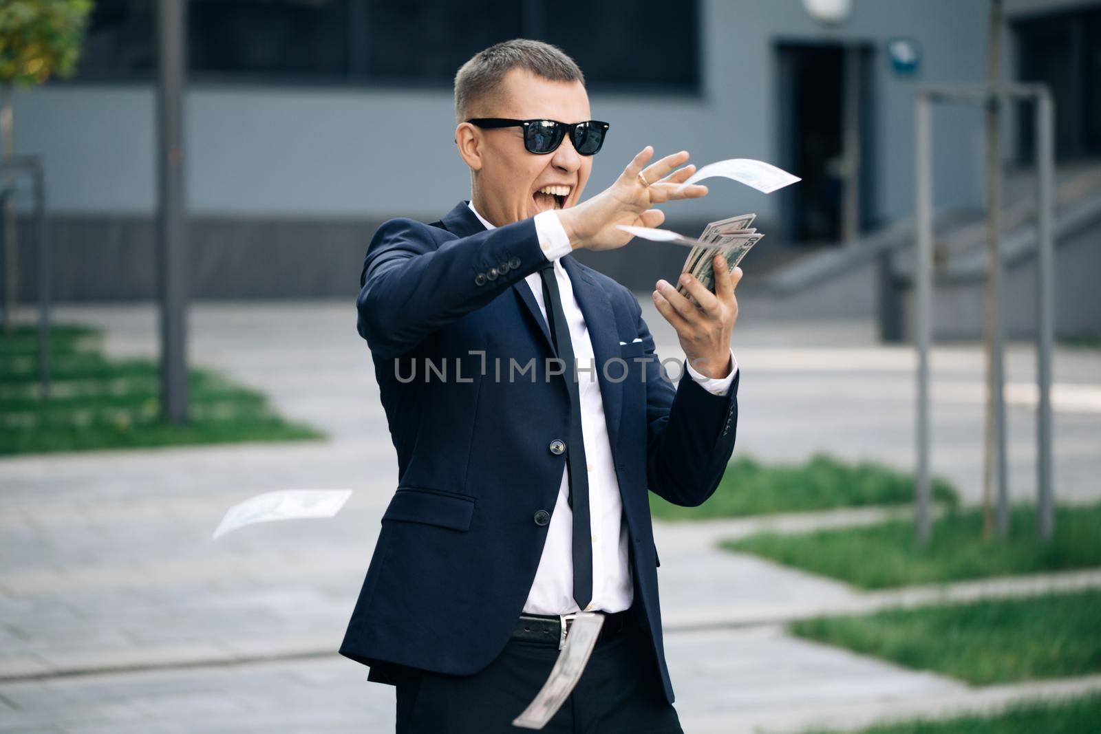 Business man with happy face scatters money. People overspend US currency. Guy is flush with dollars standing in the street near office building. Symbol of success, gain, victory.