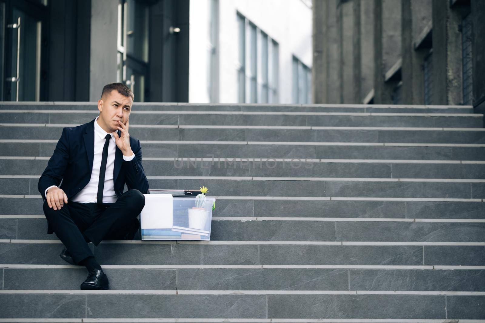 Sad businessman sitting on stairs outdoor with box of stuff as lost business. Fired man. Unemployment rate growing due pandemic. Male office worker in despair lost job.