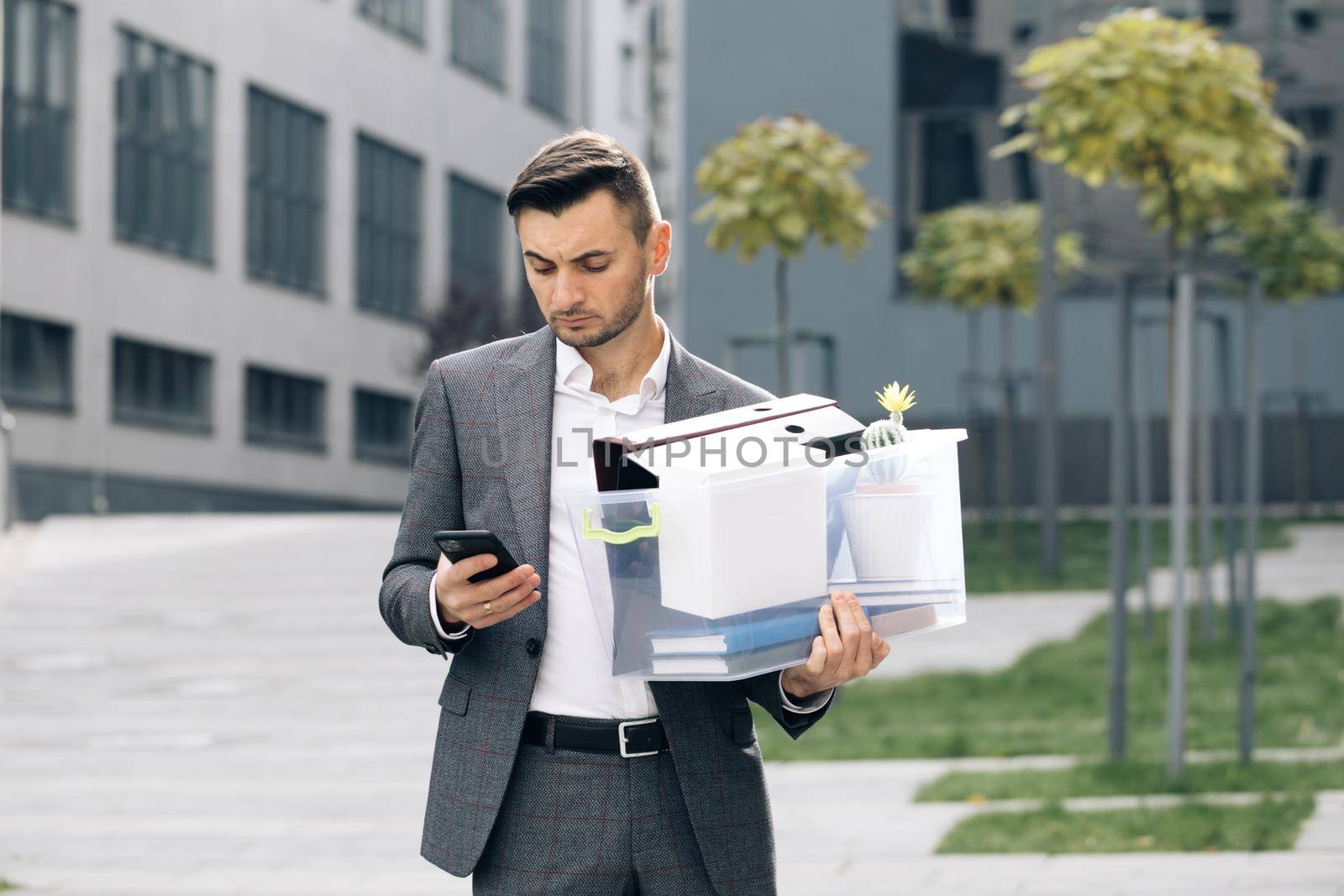 Sad young businessman uses phone texting scrolling tapping near office building. Male office worker in despair lost job. Look serious technology communication by uflypro