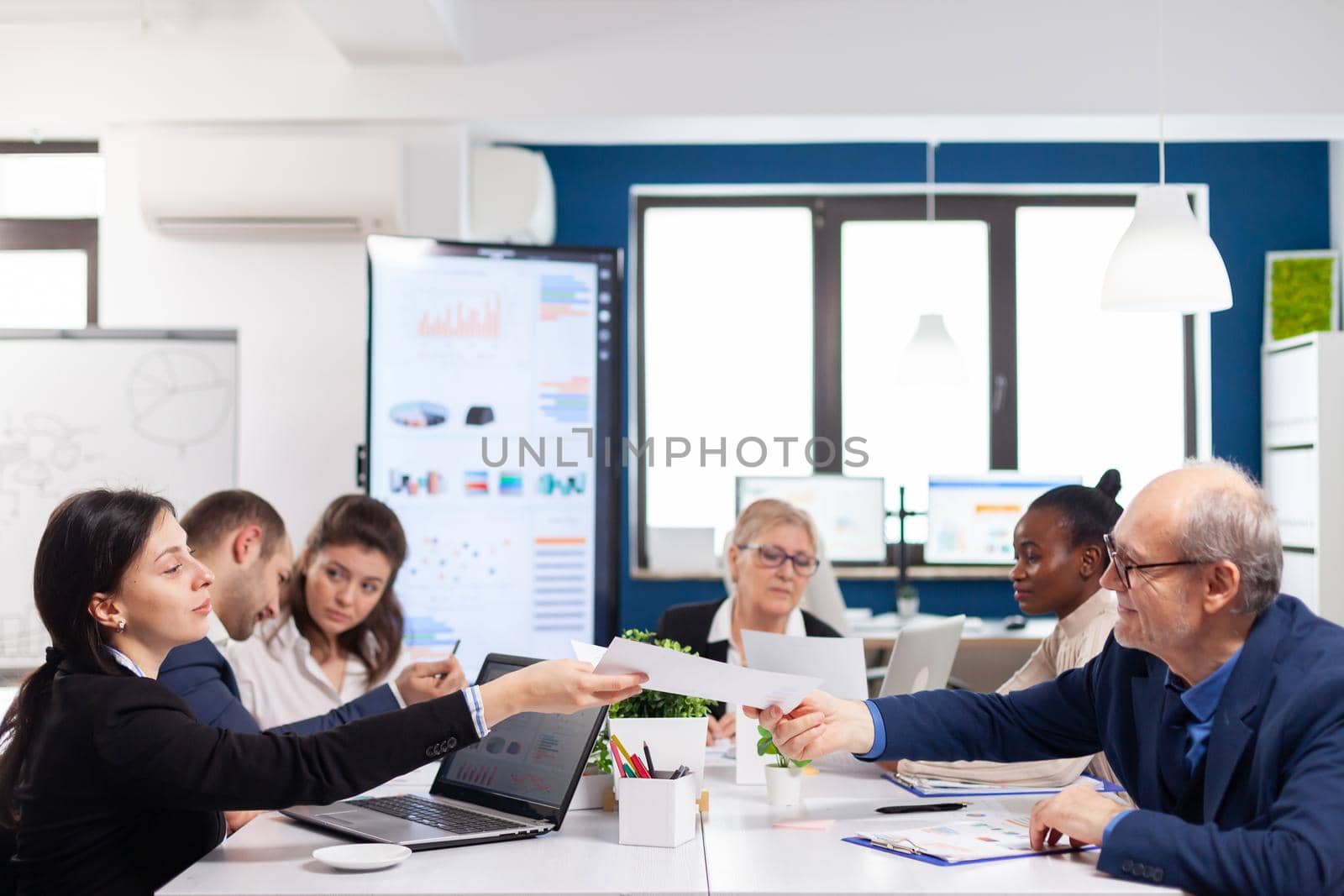 Young employee exchanging documents with senior executive in startup board room with multiethnic colleagues coworkers analysing graphs and charts in briefing corporate.