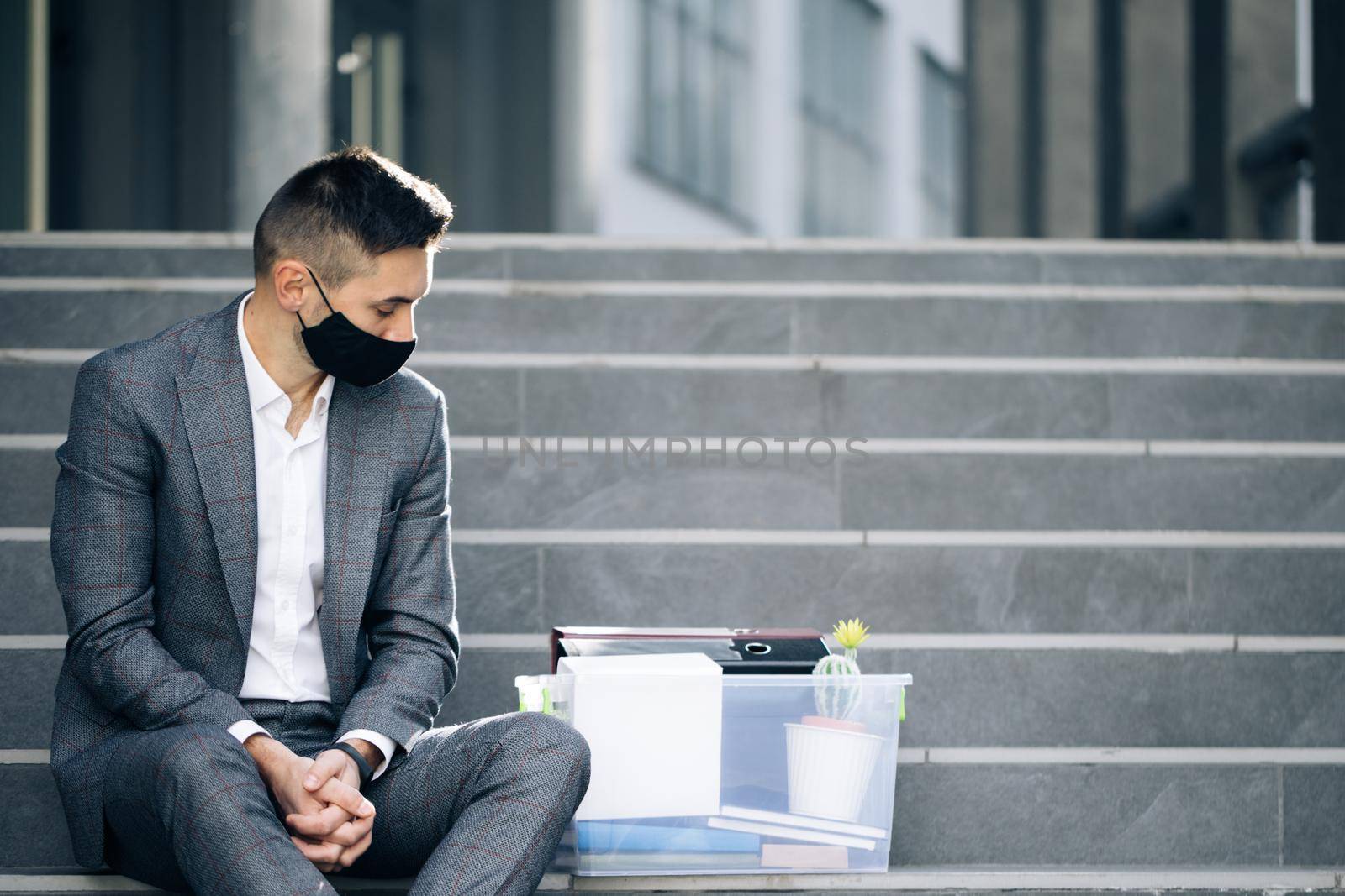 Unemployed businessman lost his business. Anxious concept. Workless man in despair. Fired male office worker in medical mask sitting on stairs in depression with box of stuff.