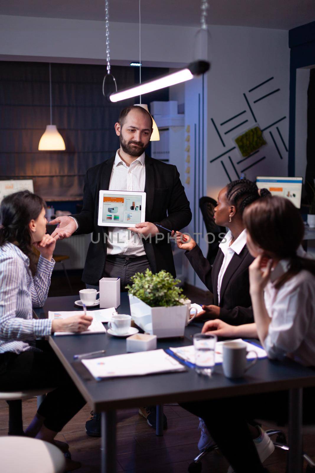 Business entrepreneur man presenting company statistics using tablet for financial presentation. Diverse multi-ethnic businesspeople brainstorming strategy working in office meeting room late at night