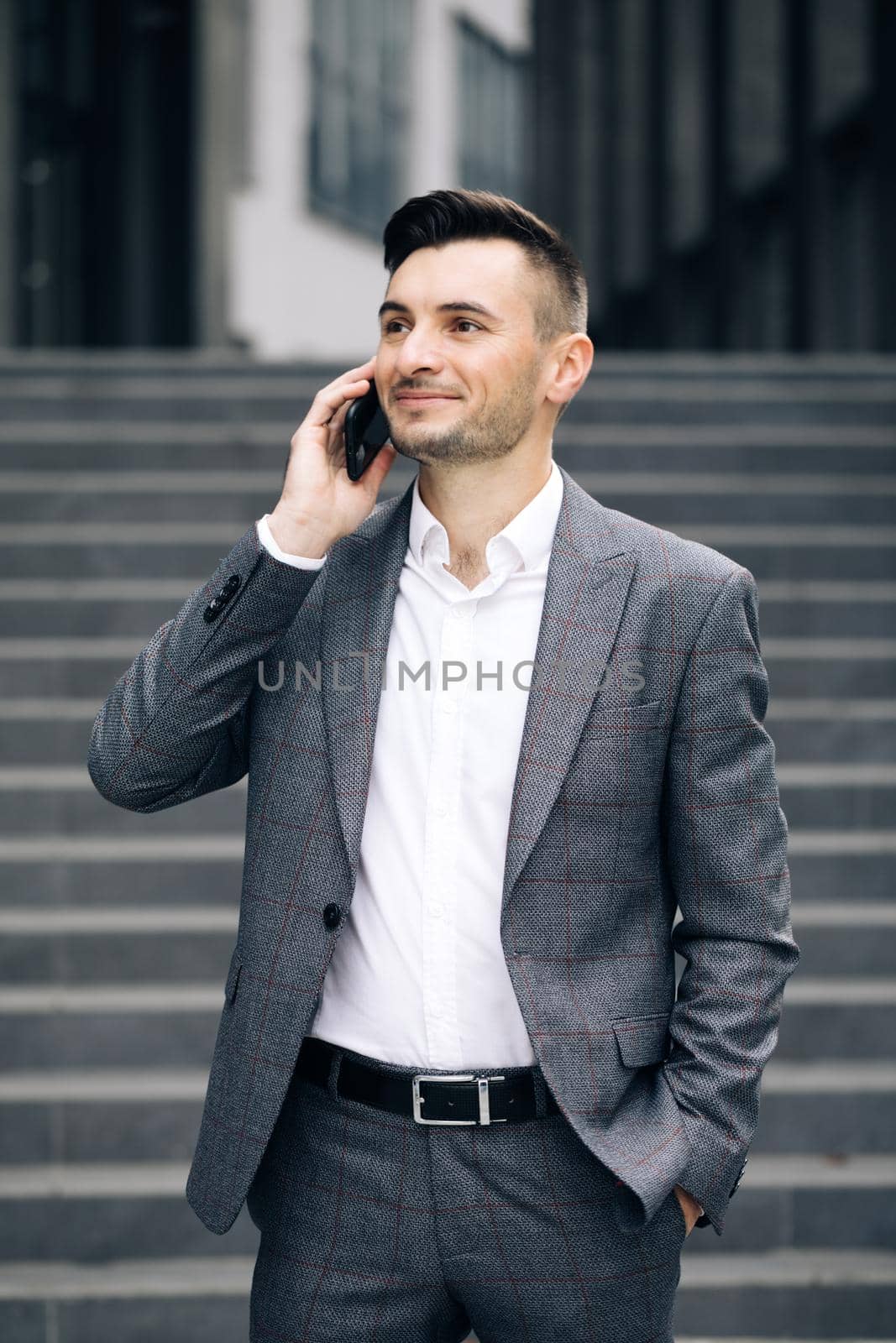 European businessman talking on mobile phone near office building background. Man is in dialogue, smiling, holding coffee cup on summer day. Vertical Screen. by uflypro