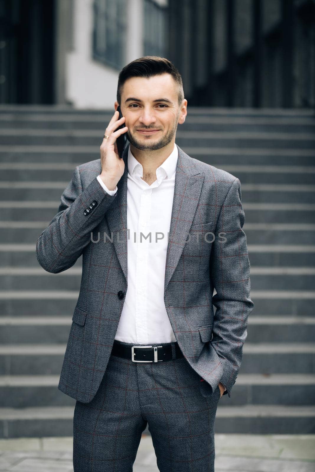 European businessman talking on mobile phone near office building background. Man is in dialogue, smiling, holding coffee cup on summer day.