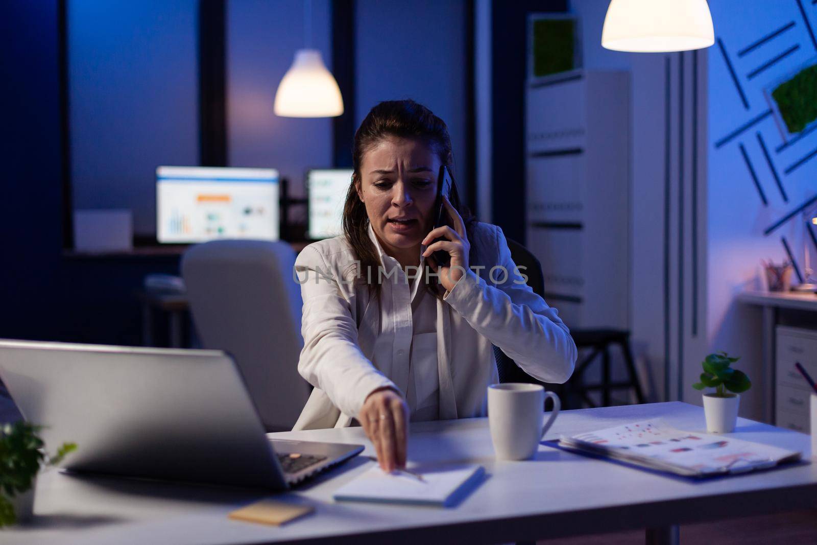 Executive woman manager speaking at phone while checking financial notes late at night. Busy focused freelancer using modern technology network wireless searching annual economic statistics