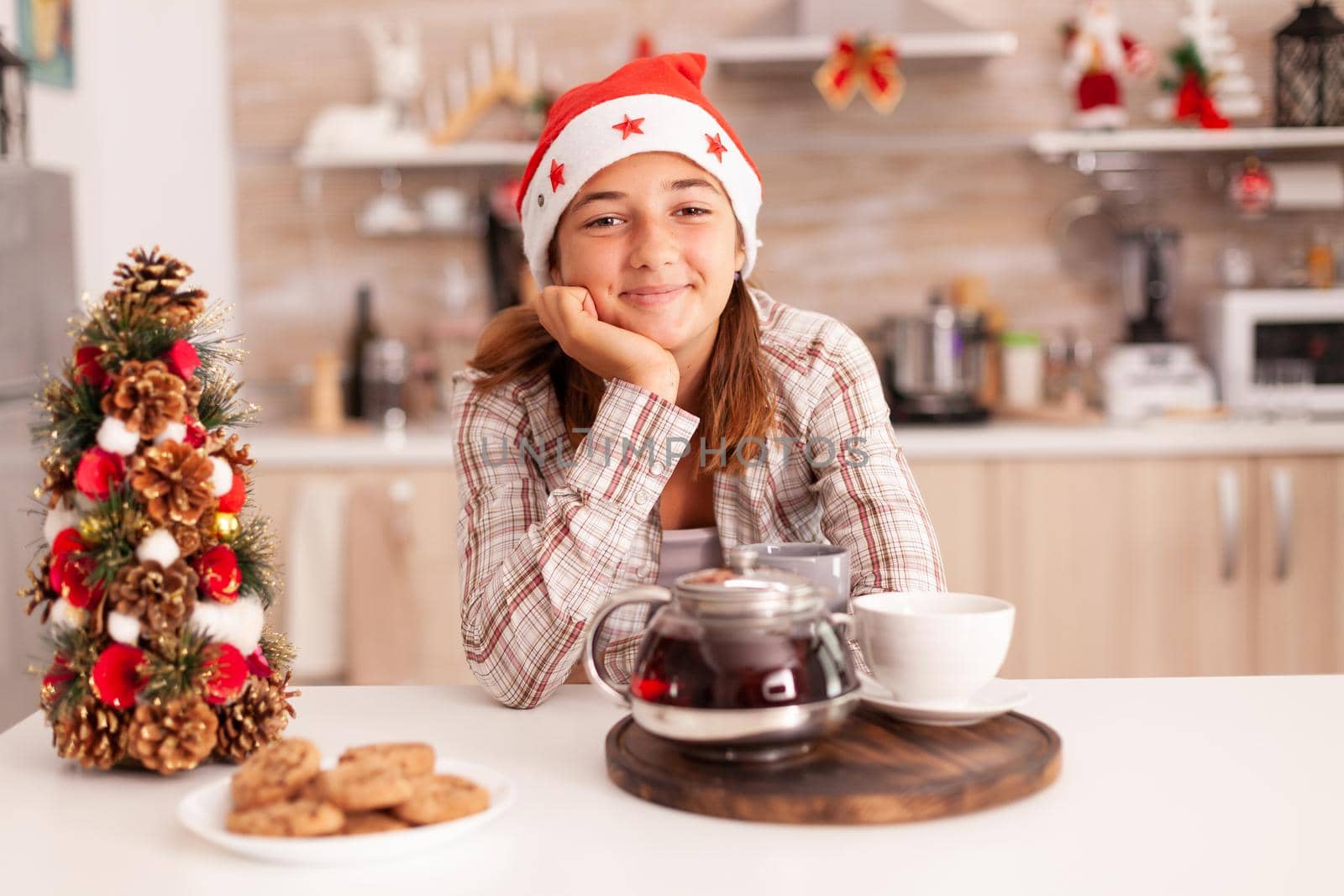 Portrait of kid with santa hat looking into camera enjoying winter holiday celebrating christmas season in xmas decorated culinary kitchen.. Baked x-mas cookies with coffee on table