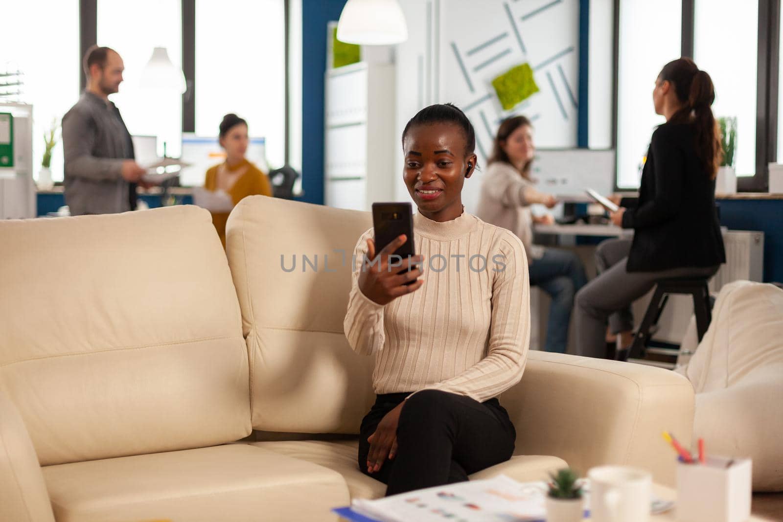 African manager woman discussing with remote colleagues on video call holding smartphone using headphones sitting on couch in business modern office. Diverse coworkers planning new financial project