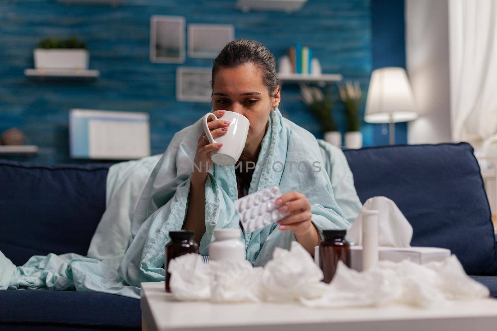 Woman with flu drinking medicine on sofa at home against cold illness disease sickness. Young caucasian person treating headache fever covid 19 virus symptoms with pills drugs