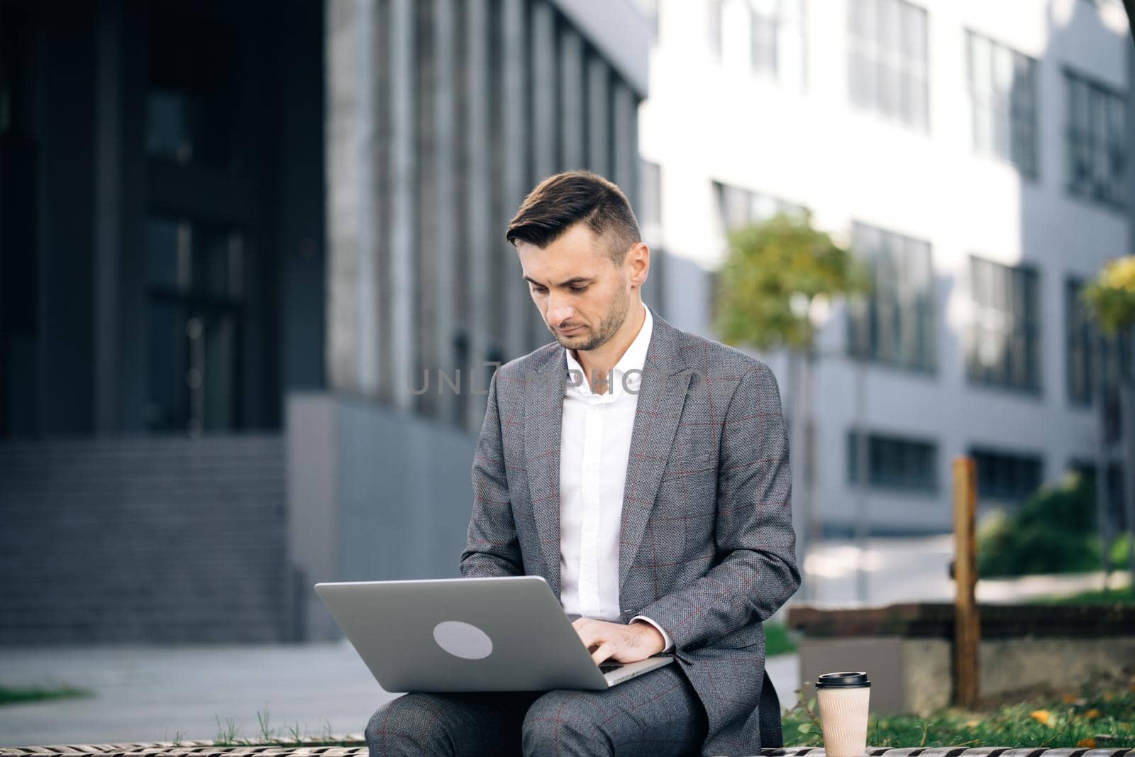 Young businessman in the city sits on bench, takes a laptop and works. Businessman typing on laptop computer outdoor. Man in suit working with laptop while sitting on bench. Remote work concept.