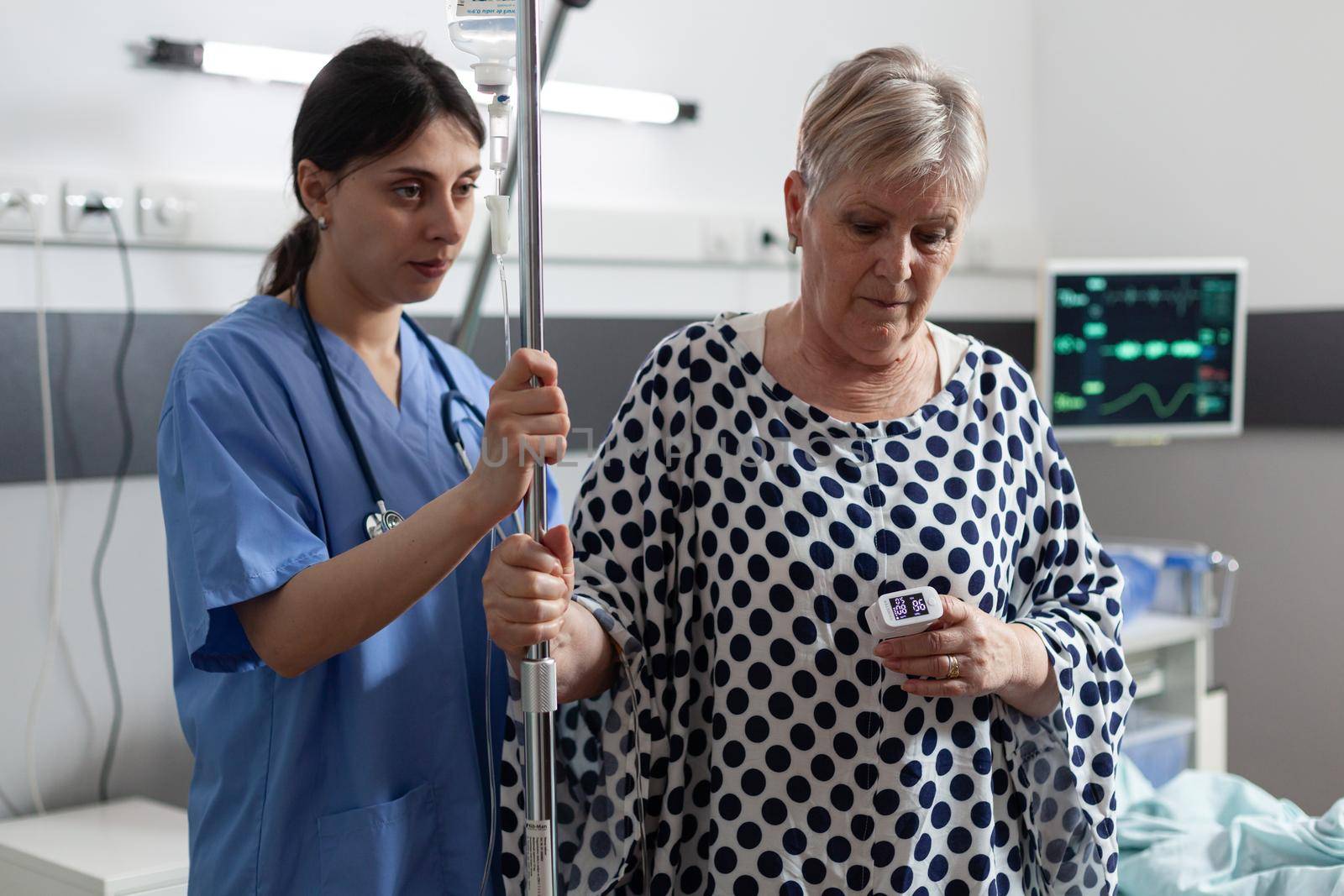 Mature old sick woman getting intravenous medicine from iv drip bag. Medical nurse helping patient walk through hospital room and showing support. Oxymeter attached on finger.