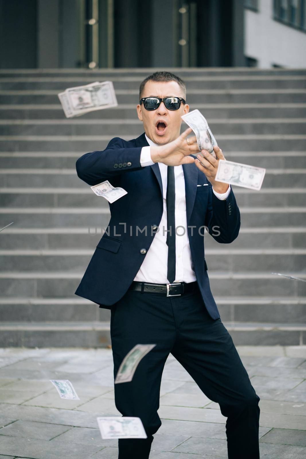 Young happy businessman scatter the dollars in the street near the office center. Money rain, falling dollars. Successful business. Funny man throws money by uflypro