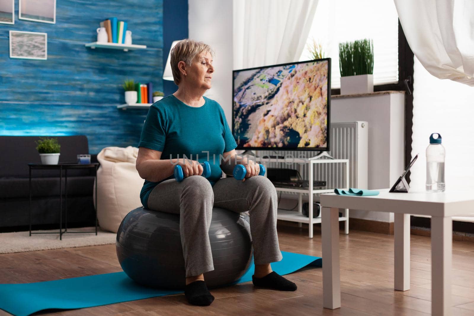 Focused senior woman doing arm exercising using workout dumbbells sitting on swiss ball by DCStudio