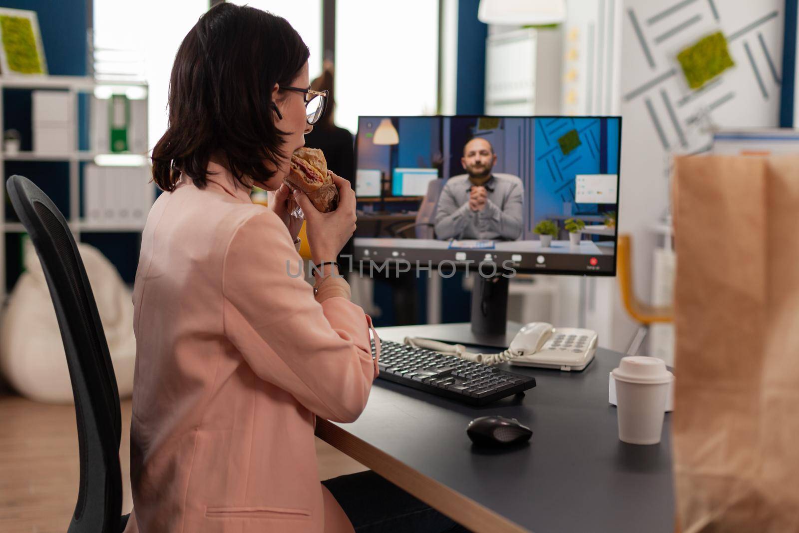 Entrepreneur woman sitting at desk in company office eating sandwich during online videocall conference by DCStudio