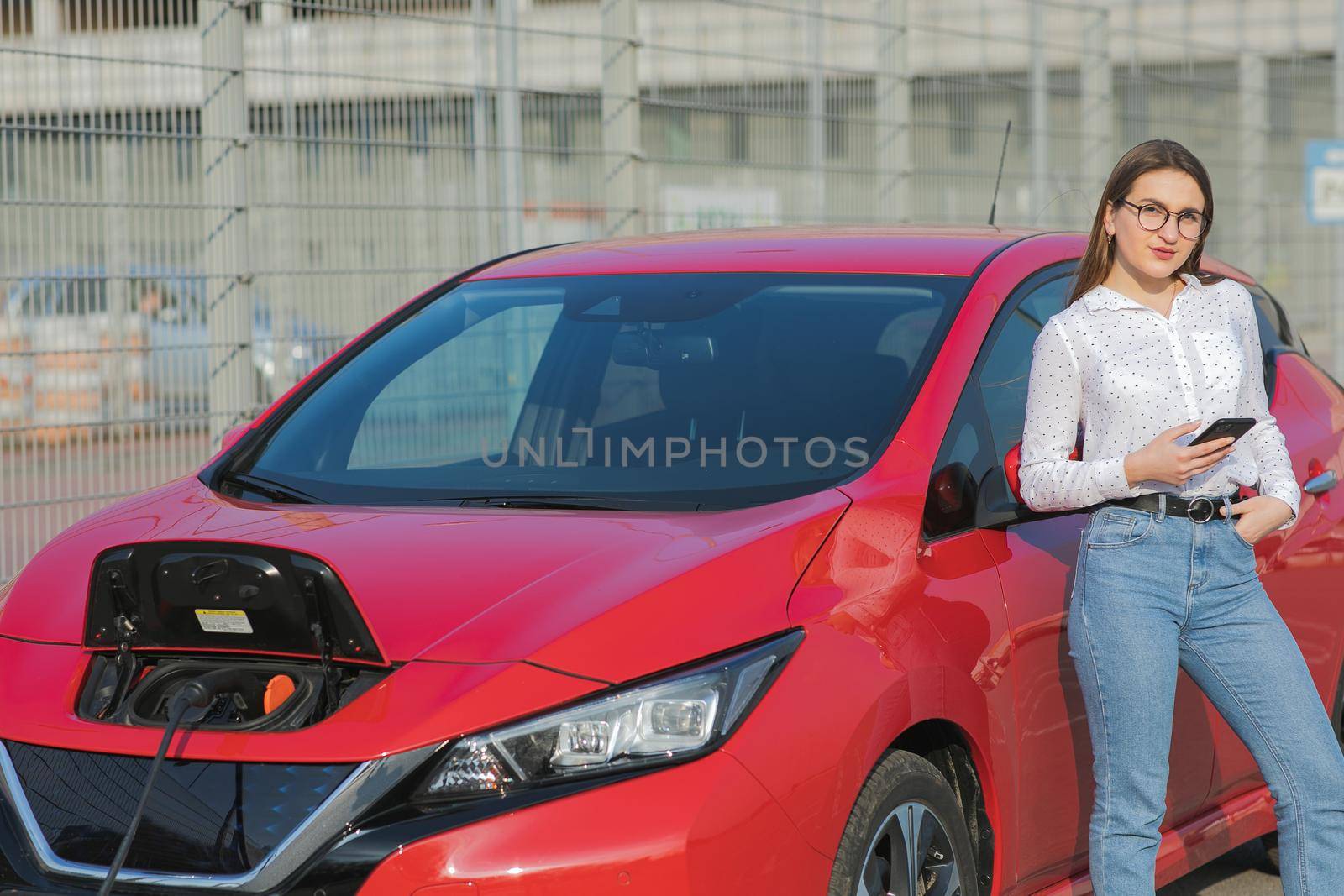 Girl stands with phone near her red electric car and waits when vehicle will charged. Connecting the Charger Plug of an Electric Car
