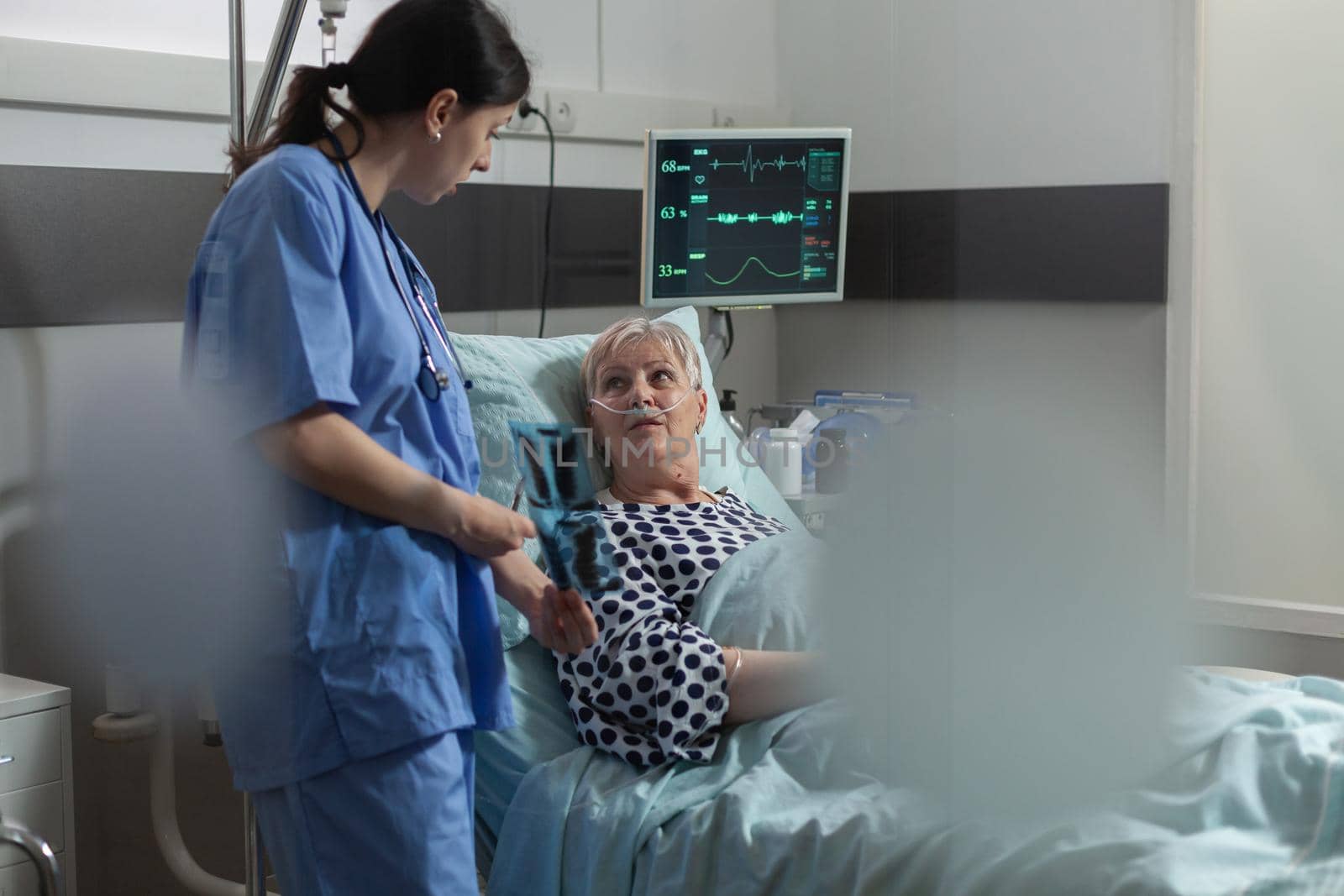 Elderly patient in radiology hospital room laying in bed listening nurse dicussing about chest x-ray radiography for respiratory diagnosis. Breathing with help from oxygen mask.