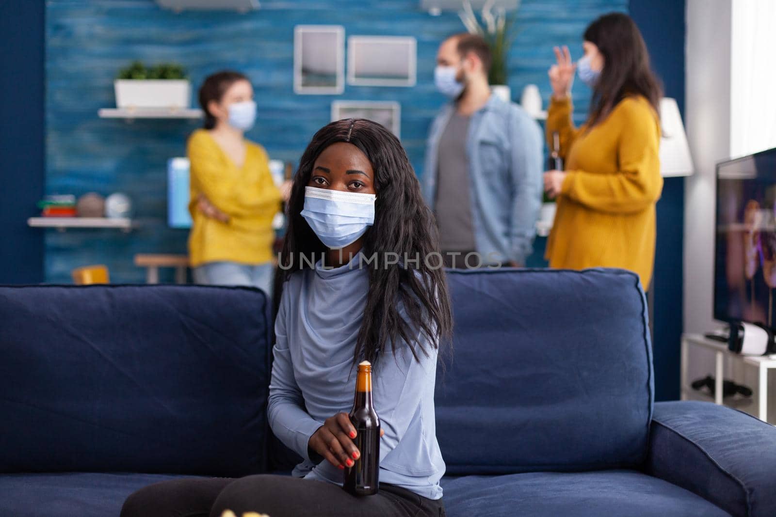 African woman keeping social distancing wearing face mask while meeting with friends to prevent spread of coronavirus holding beer bottle looking at camera sitting on couch. Conceptual image.