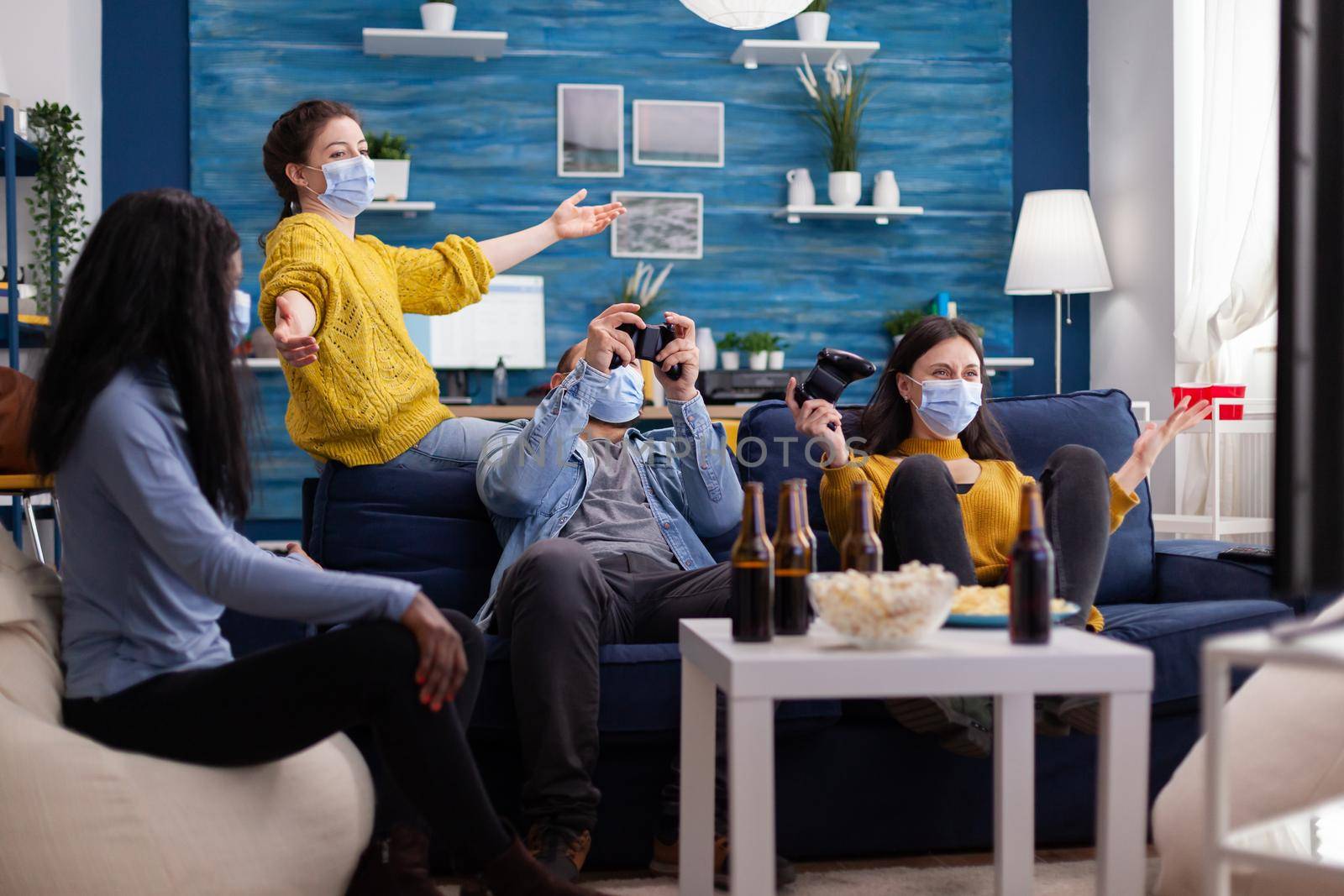 Group of multiethnic friends having a gaming competition in apartment home living room using wireless joystick sitting on sofa keeping social distancing wearing face mask in the course of coronavirus global outbreak.