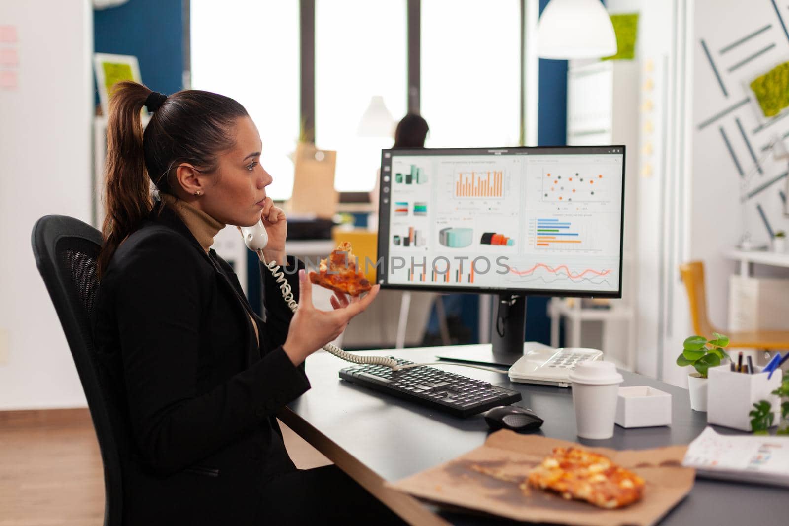 Closeup of businesswoman sitting at desk in front of computer eating pizza slice by DCStudio