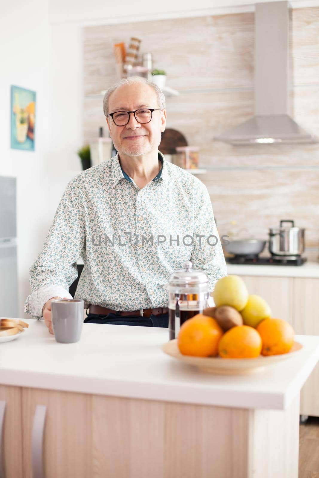 Elderly man in kitchen smiling looking at camera holding hot coffee cup. Portrait of relaxed older person in the morning, enjoying fresh warm drink. Healthy smiling adult face