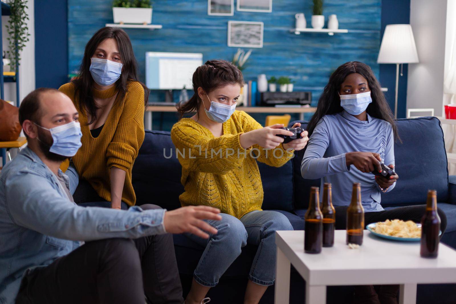 Concentrated multiethnic friends trying to win at video games during social pandemic wearing face mask keeping distancing not to spread corona sitting on couch in living room.