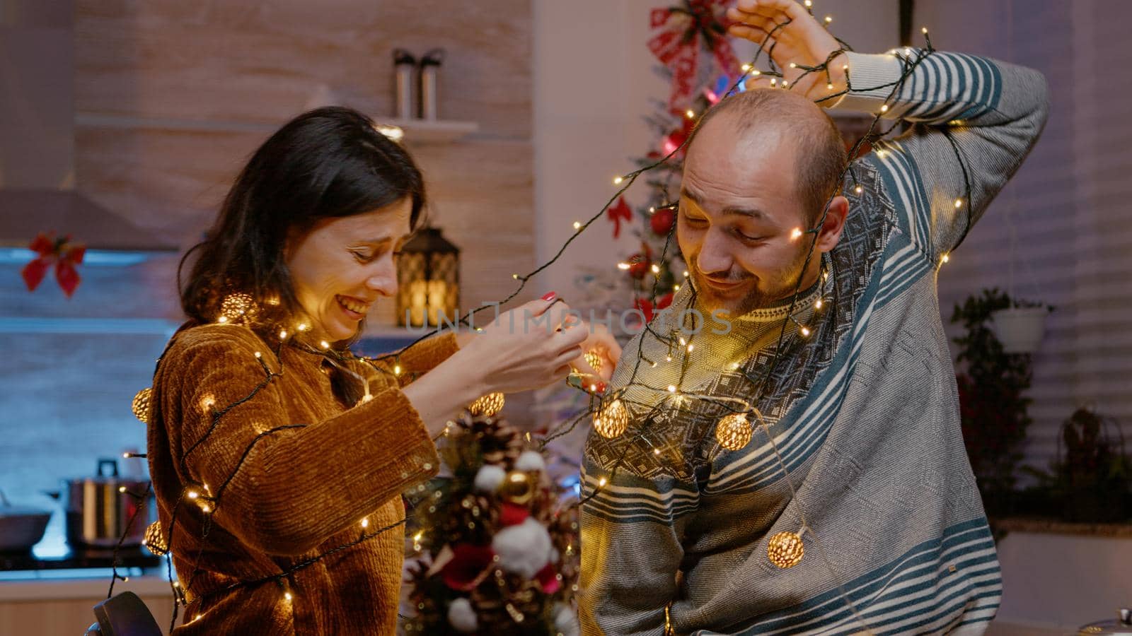 Couple laughing getting tangled in string of festive lights while decorating kitchen for christmas eve celebration. People knotted in garland of illuminated bulbs and twinkle lights