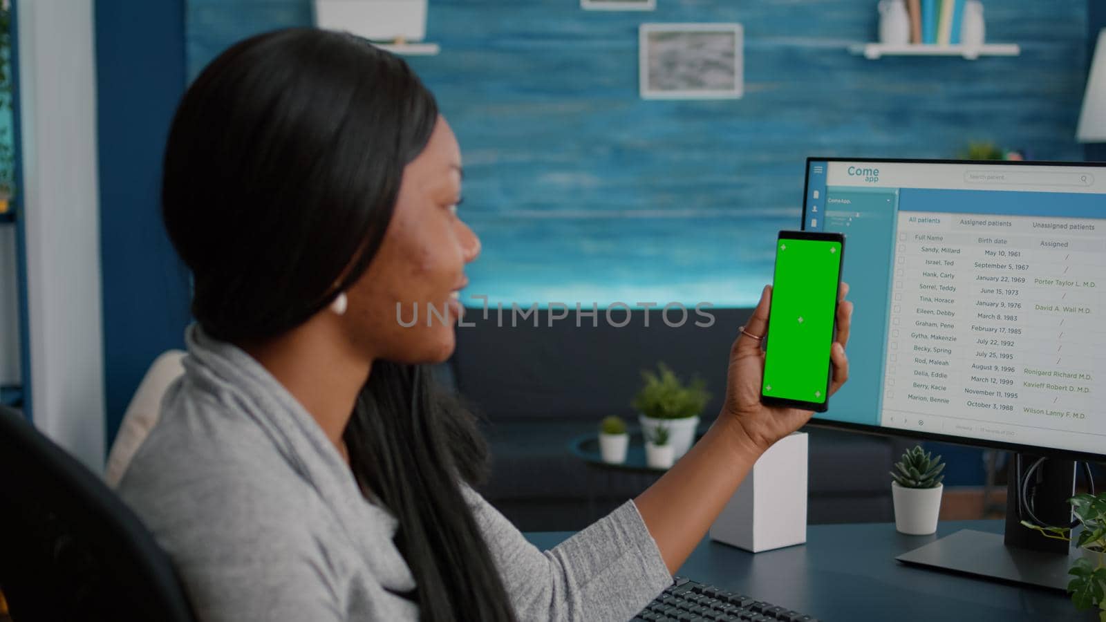 Student with dark skin holding phone with mock up green screen chroma key looking at isolated display sitting at desk table in living room. Woman texting message working at social network app