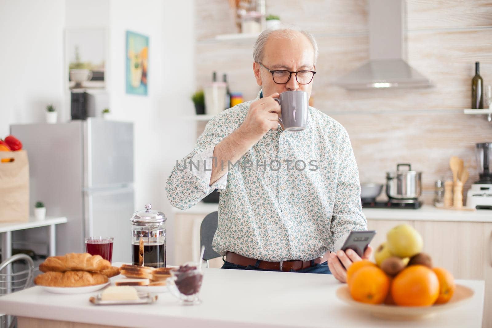 Senior man drinking coffee during breakfast in ktichen and using smartphone.