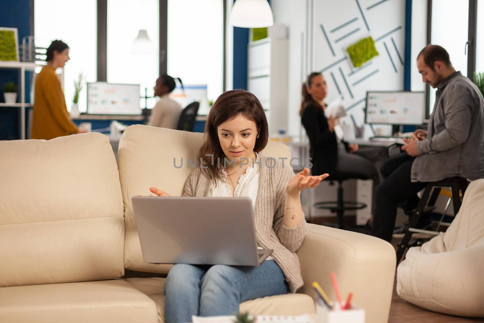 Manager woman sitting on couch holding laptop and talking on video call during virtual conference working in business modern office. Diverse coworkers planning new financial project in modern company