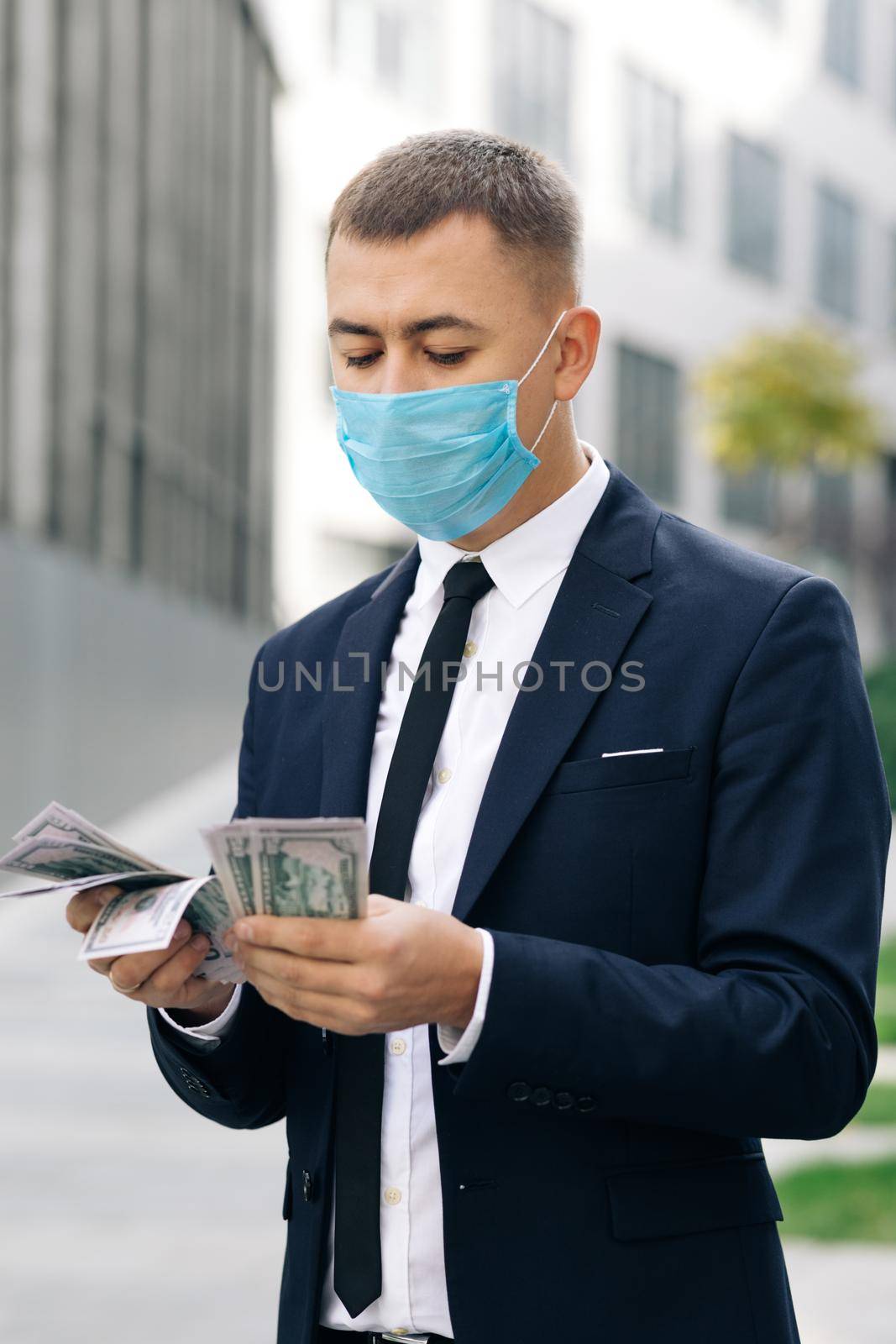Business Man in Protective Face Mask Counts Cash. Quarantine financial crisis. Young Handsome Rich Man Wearing Stylish Suit Counting Money Standing in the street near Office Building by uflypro