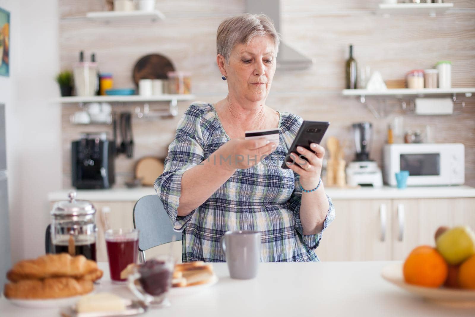 Retired old woman using credit card for online shopping in kitchen during breakfast. Senior elderly person using internet payment home bank buying with modern technology