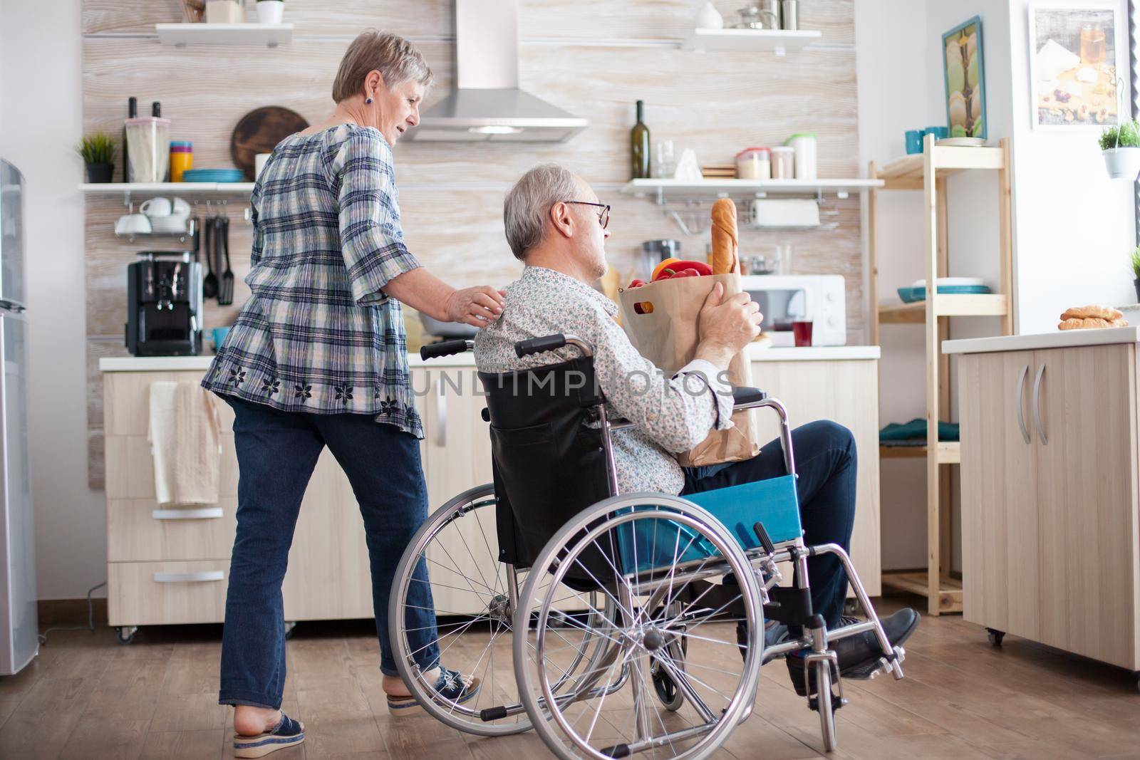 Senior woman taking grocery paper bag from handicapped husband in wheelchair. Mature people with fresh vegetables from market. Living with disabled person with walking disabilities