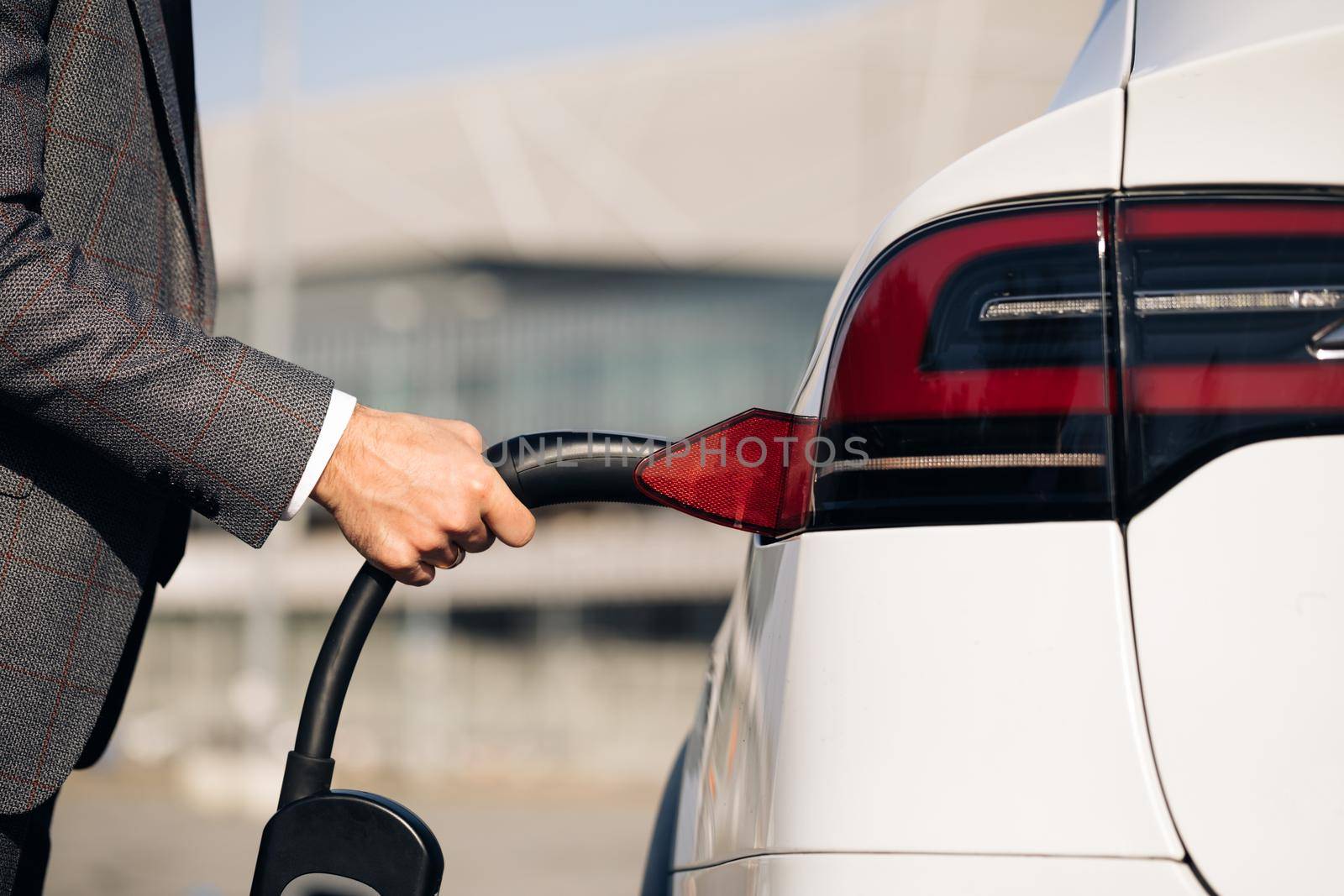 Unrecognizable man plugging in electric car at electroic car charging station. Luxury white electrical car recharging. Environmentally conscious male charging electric vehicle.