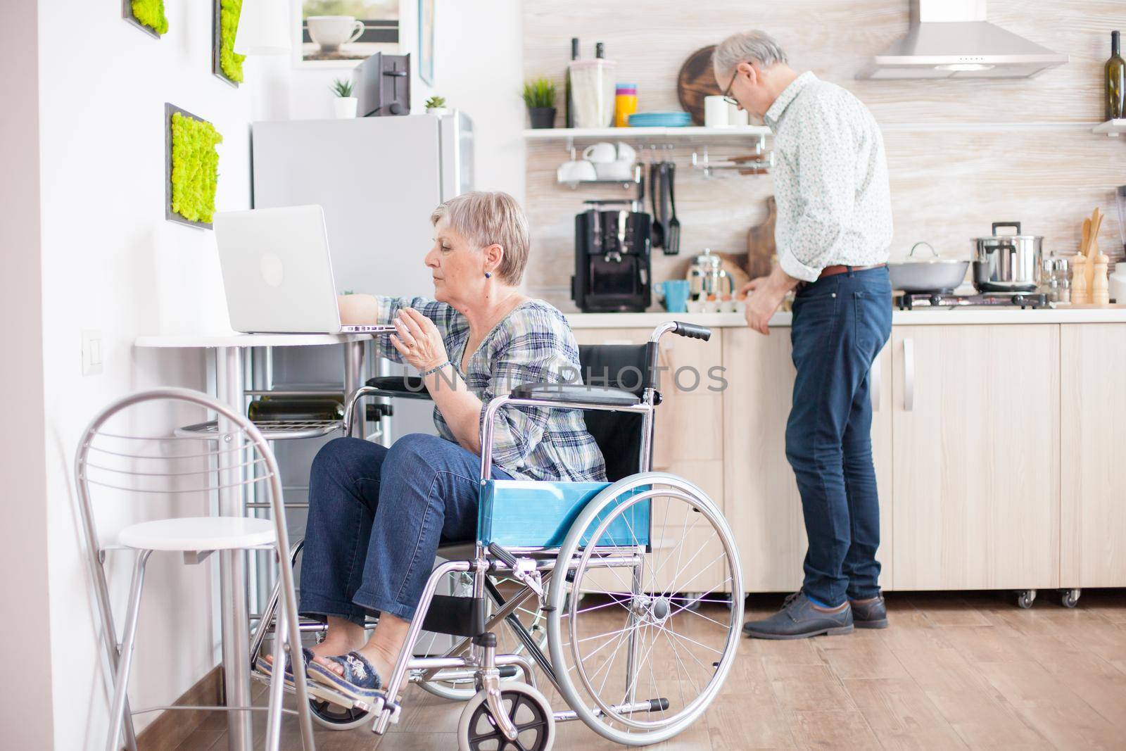 Disabled old woman in wheelchair working on laptop in kitchen. Paralyzed handicapped old elderly person using modern communication online internet web techonolgy.