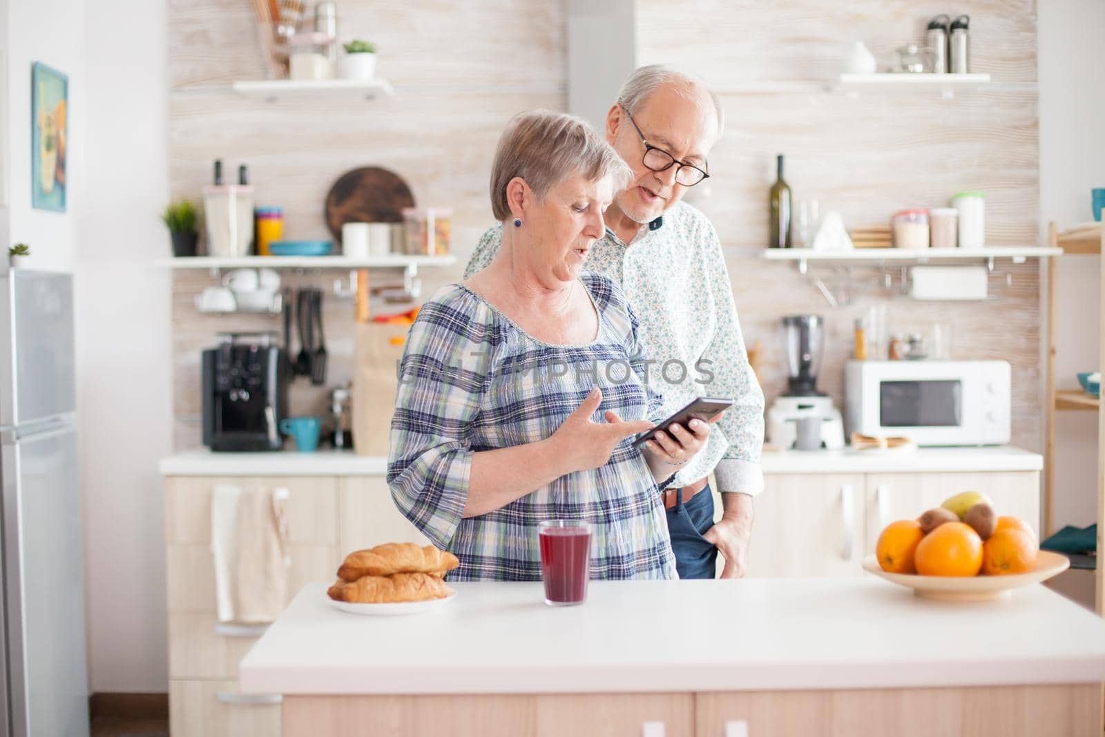 Caucasian senior wife and husband in kitchen during breakfast using smartphone technology and internet conection. Leisure retired woman and man smiling in domicile, relaxing.