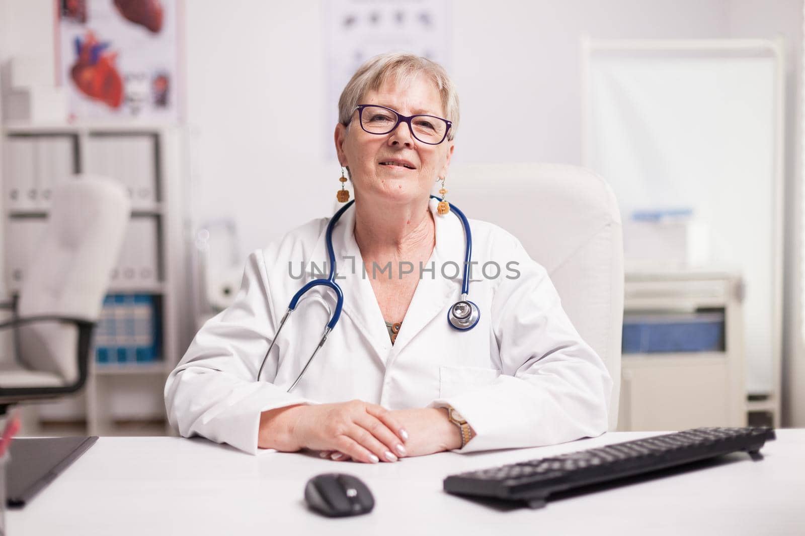 Senior doctor with stethoscope wearing white coat smiling to camera in hospital cabinet.