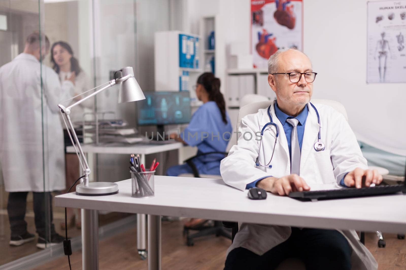 Senior specialist doctor in clinic office using computer to write patient health report wearing white coat while young medic is taking information about sick person on hospital corridor.