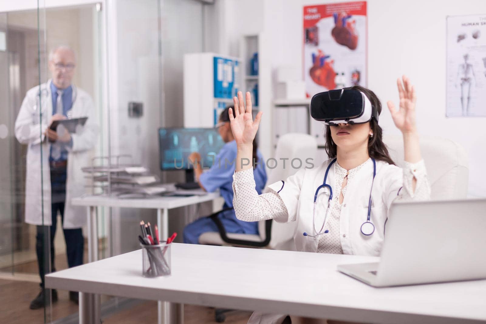 Young doctor using augmented reality goggles in hospital office while senior medic is taking notes on clipboard in clinic corridor and nurse working on computer.