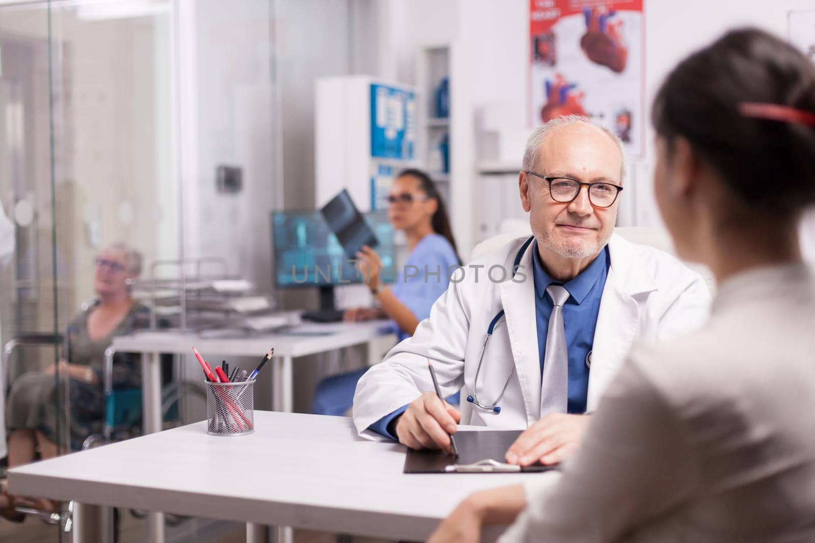 Senior doctor talking with patient about her illness in hospital office. Nurse holding x-ray image in the background. Elderly aged woman in wheelchair.