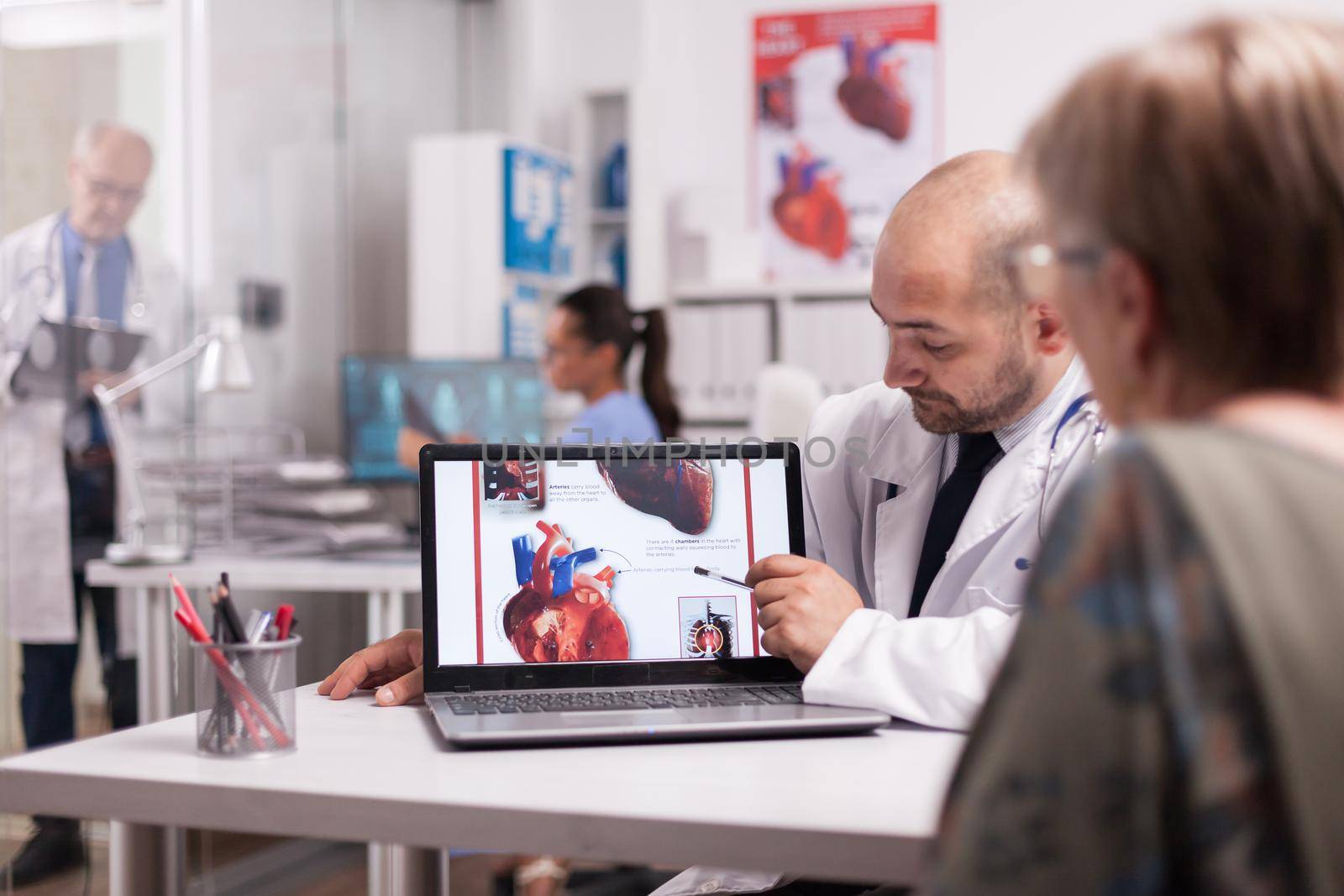 Cardiologist in white coat pointing at heart on laptop screen in hospital office and talking with old woman about treatment for her illness. Mature doctor writing prescription on clipboard in clinic corridor.