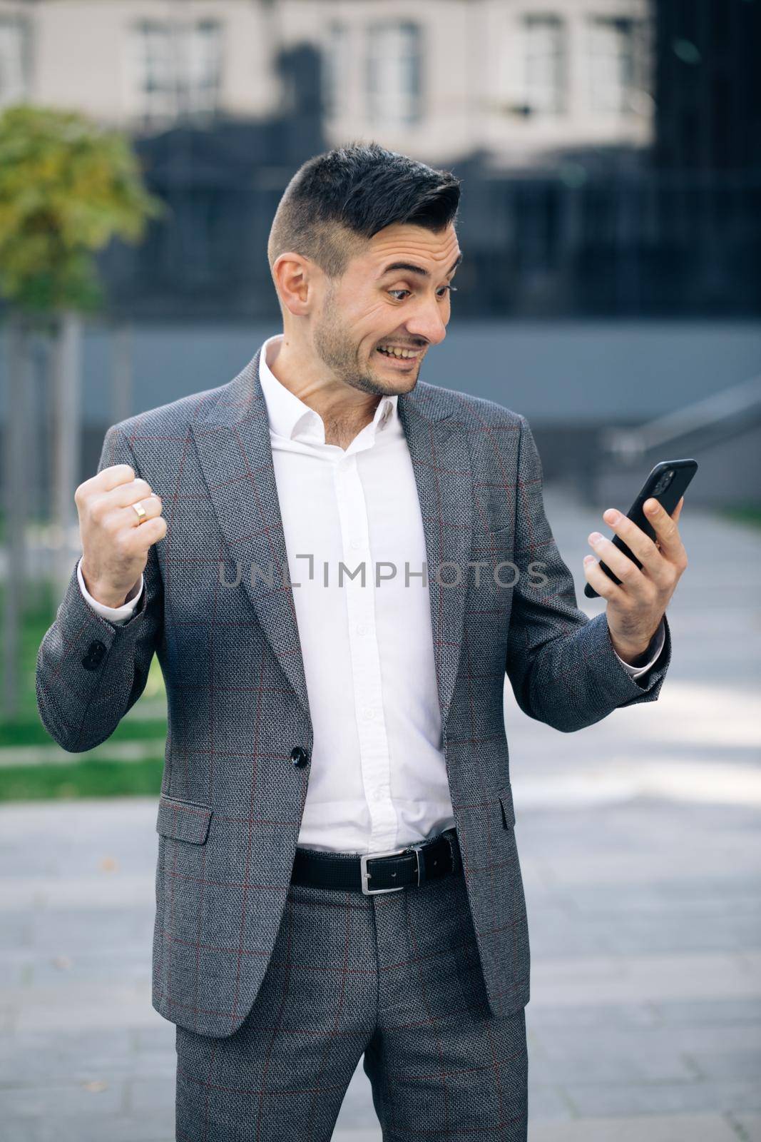 Successful businessman celebrating win with mobile phone outside. Surprised man looking mobile phone outside. Portrait of excited man reading good news at smartphone outdoors.