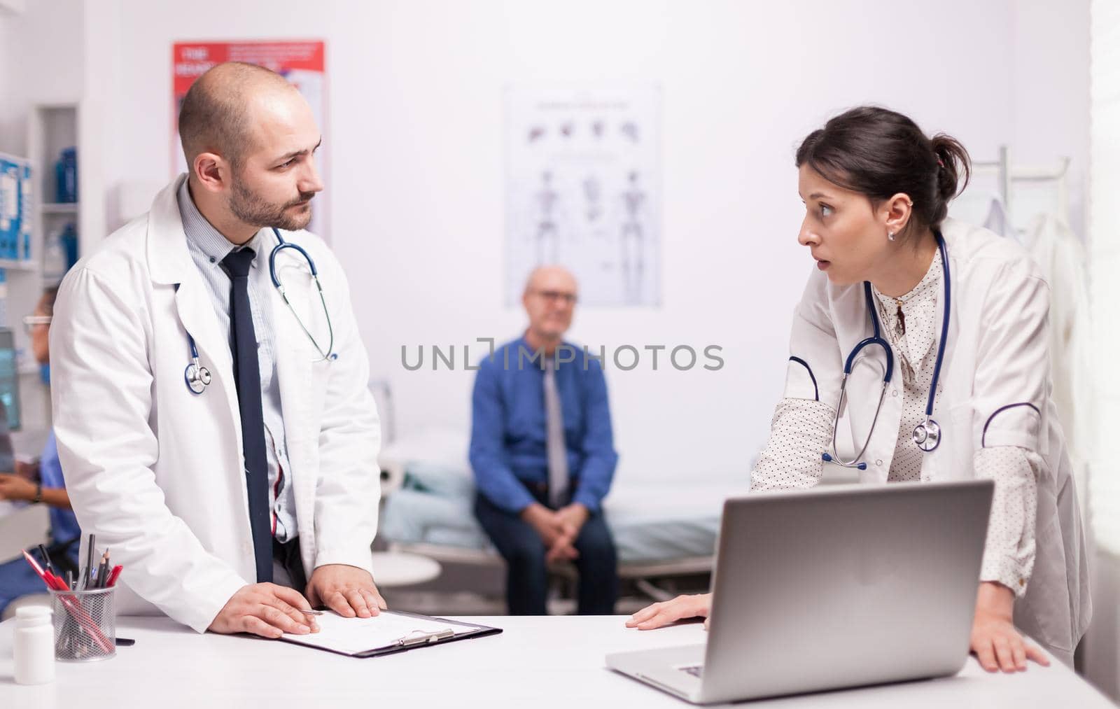 Doctors looking at each other wearing white coat and stethoscope checking senior patient diagnosis on laptop in hospital office.