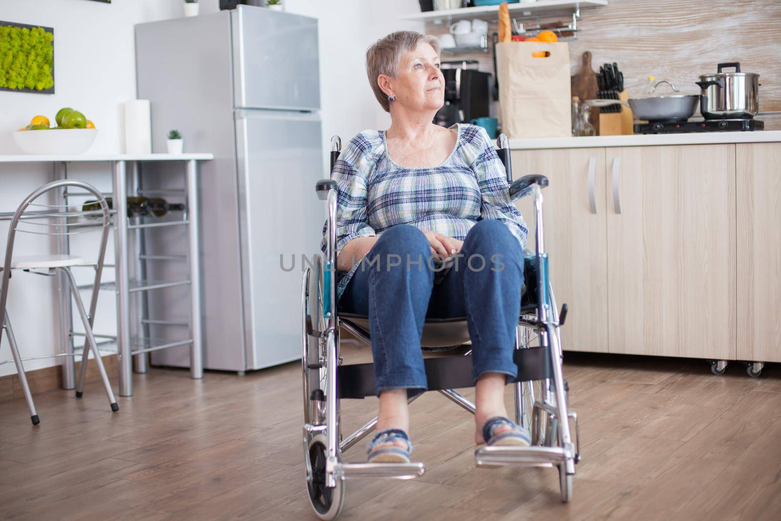 Handicapped senior woman in wheelchair standing alone in kitchen looking throug window. Pensive thoughtful lonely woman in solitude. Elderly disabled pensioner after injury and rehab, paralysis and disability depressed invalid full of sorrow, worry and sad face. Retirement period for old people