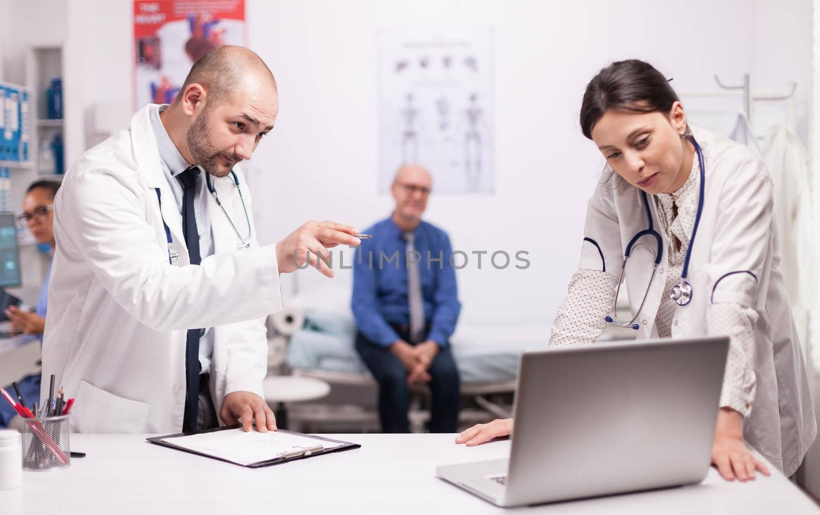 Team of doctors checking patient diagnosis on laptop while he is sitting worried on hospital bed. Senior man during consultation. Medics with stethoscope.