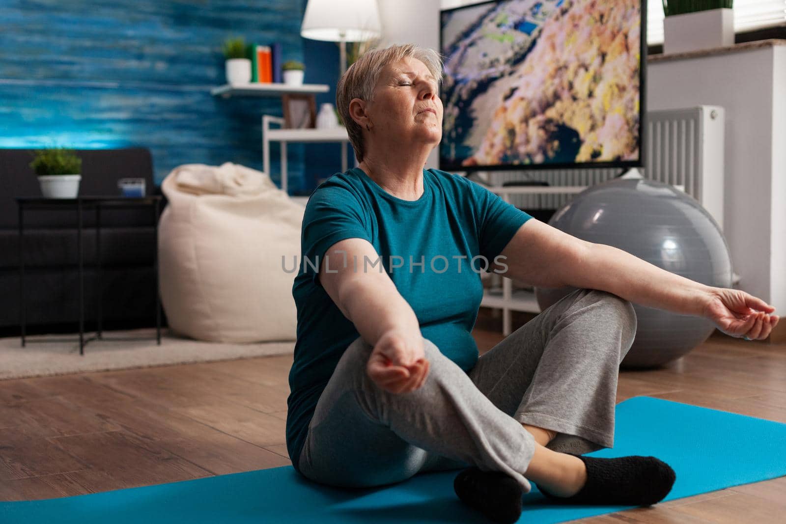 Senior woman sitting comfortable in lotus position on yoga mat with closed eyes meditating. Peaceful pensioner practicing fitness body exercise during meditation workout in living room
